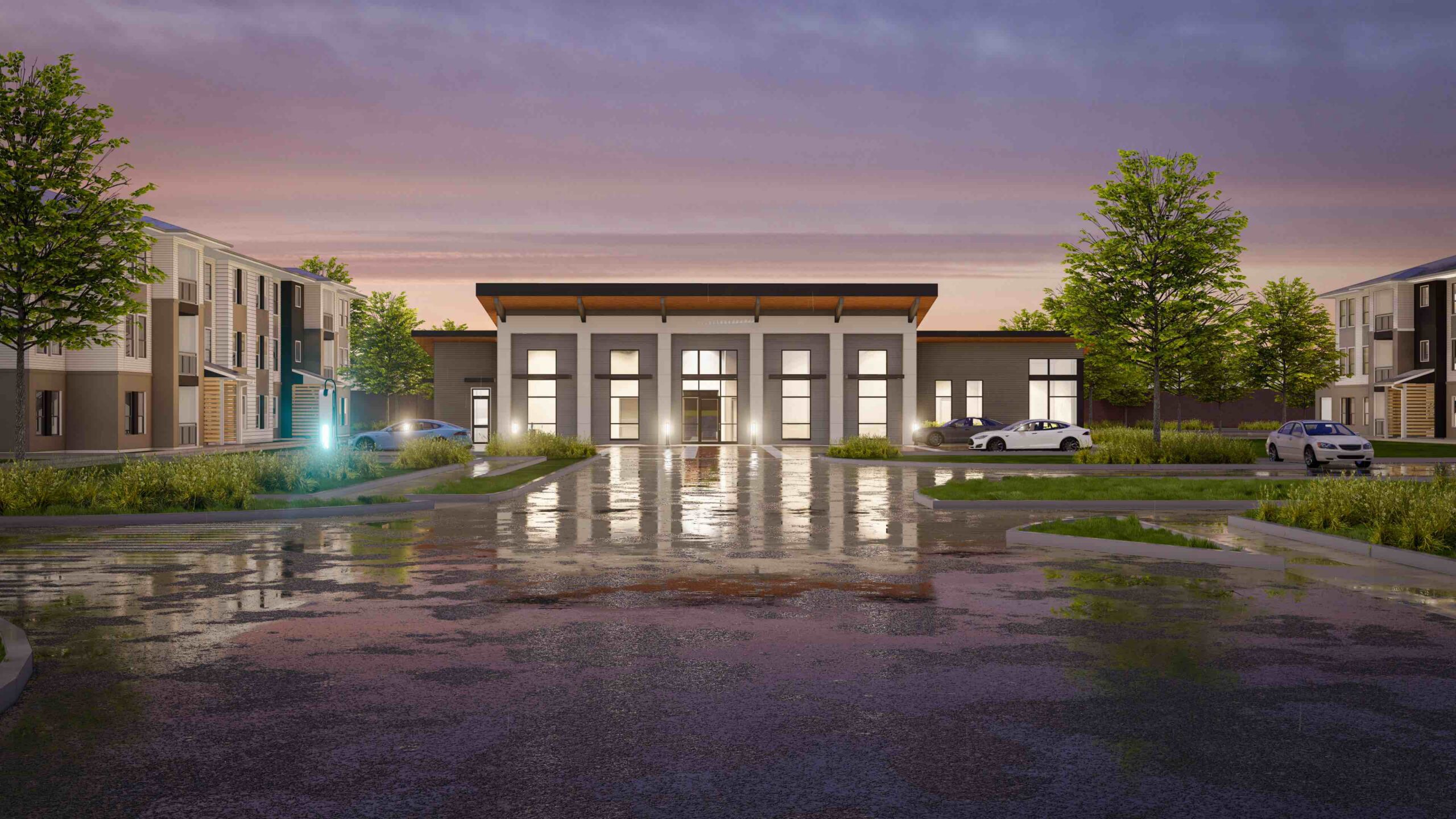 Modern apartment complex at dusk with wet pavement reflecting light, cars parked on either side, and a central building with large windows and a flat roof. Trees are visible around the area.