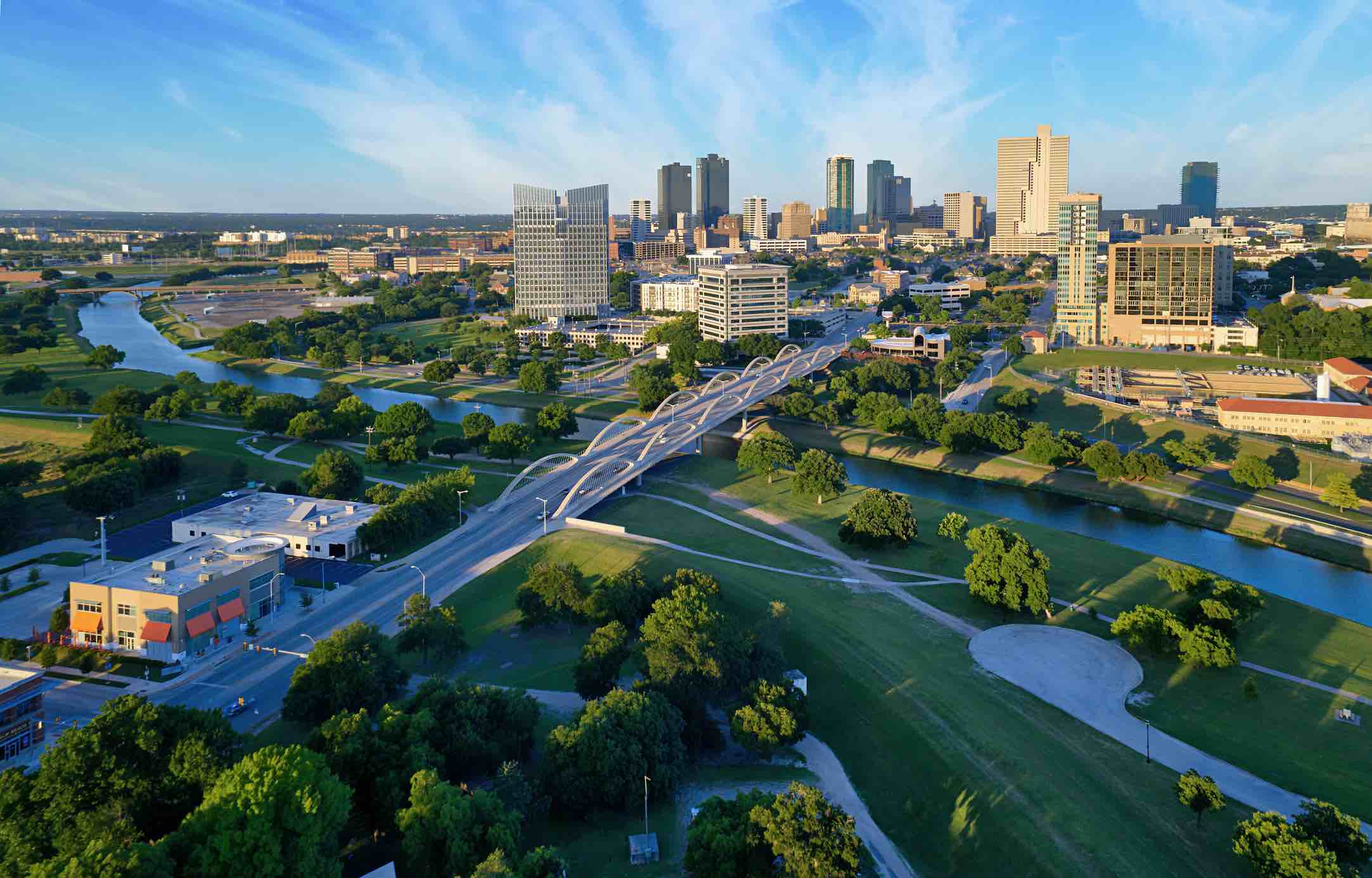 Aerial view of the Fort Worth TX skyline with modern buildings, a river, multiple bridges, and green parks under a clear blue sky, showcasing the vibrant appeal of apartments in Fort Worth.