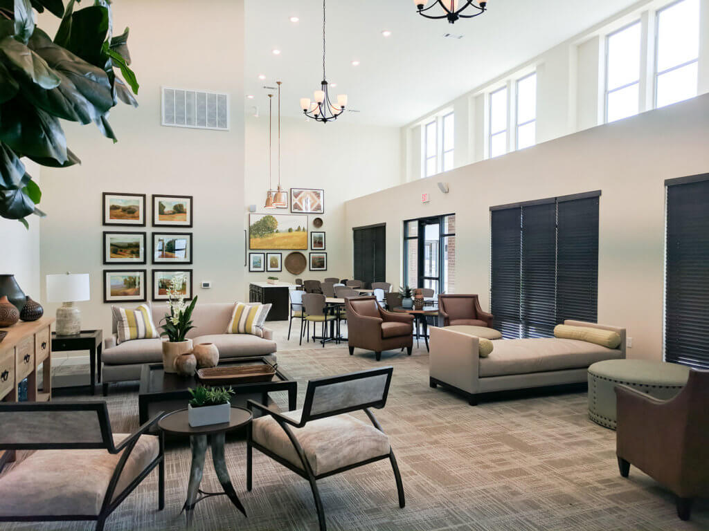 Modern lounge area with sofas, armchairs, and tables in neutral tones at Columbia Renaissance Square, featuring wall art, large windows with blinds, and a dining space in the background under high ceilings with pendant lights.