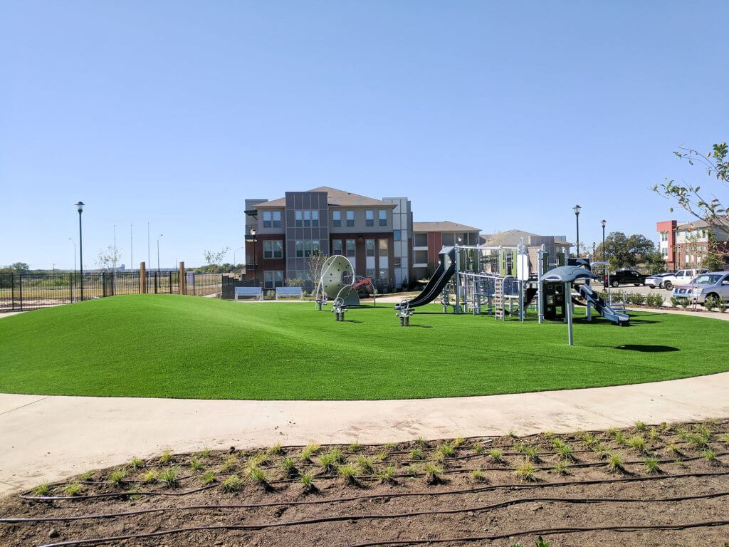 A small playground with slides and climbing equipment sits on artificial turf, surrounded by a sidewalk and the modern apartment buildings of Columbia Renaissance Square under a clear sky.