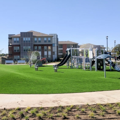 Playground with slides and climbing equipment on artificial grass at Columbia Renaissance Square, surrounded by apartment buildings under a clear blue sky.