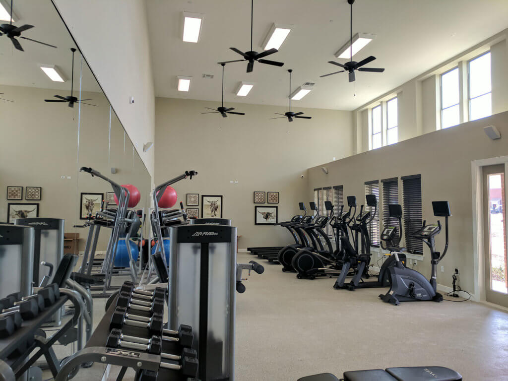 A modern gym at Columbia Renaissance Square features exercise machines, free weights, large mirrors, ceiling fans, and natural light streaming through tall windows.