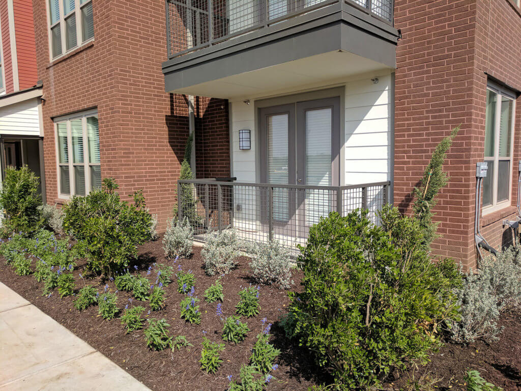 Ground-level apartment patio at Columbia Renaissance Square with metal railing, double doors, and a landscaped garden bed featuring shrubs and small plants in front of a red brick building.
