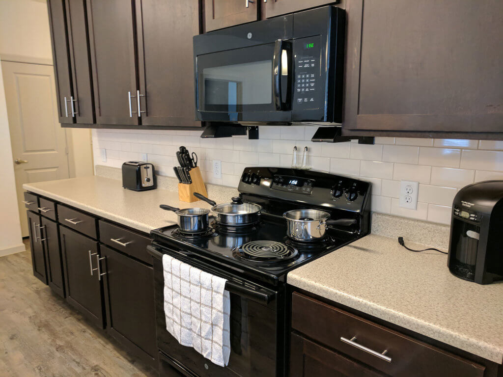 Modern kitchen at Columbia Renaissance Square with dark cabinets, a microwave, stove with three pots, a coffee maker, toaster, and a knife block on a light countertop; towel hanging on the oven door.