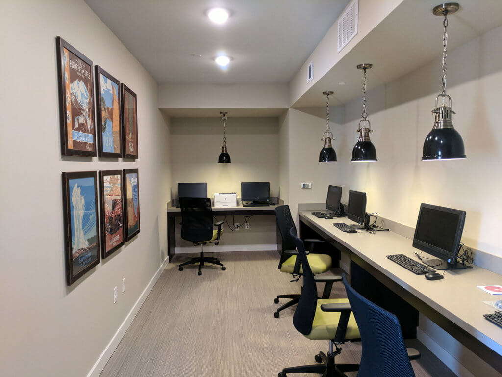 A small computer lab at Columbia Renaissance Square features four computers, a printer, framed posters on the wall, pendant lights above, and office chairs along two work surfaces.