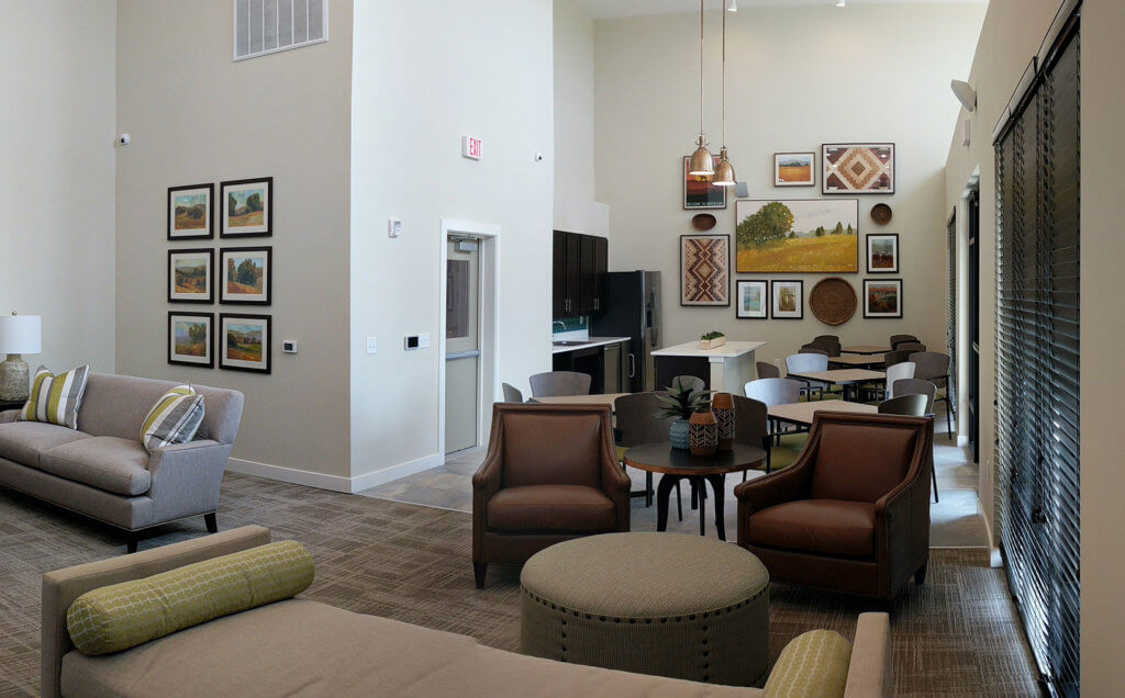 Modern lounge area at Columbia Renaissance Square featuring sofas, armchairs, a round table, wall art, and large windows with blinds; neutral tones and bright lighting create an inviting ambiance.